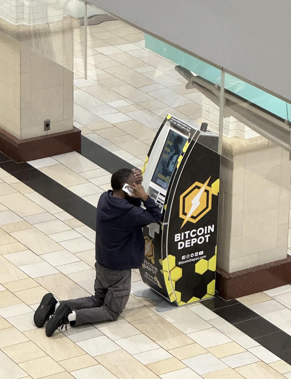 A man kneels in front of a Bitcoin ATM, speaking on the phone and apparently in distress.
