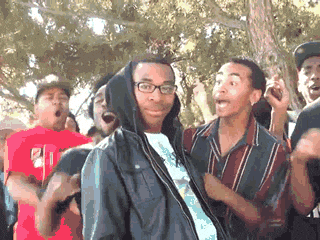 An animated GIF of a young man in glasses and a black jacket standing calmly while a crowd around him reacts excitedly, cheering and gesturing outdoors.